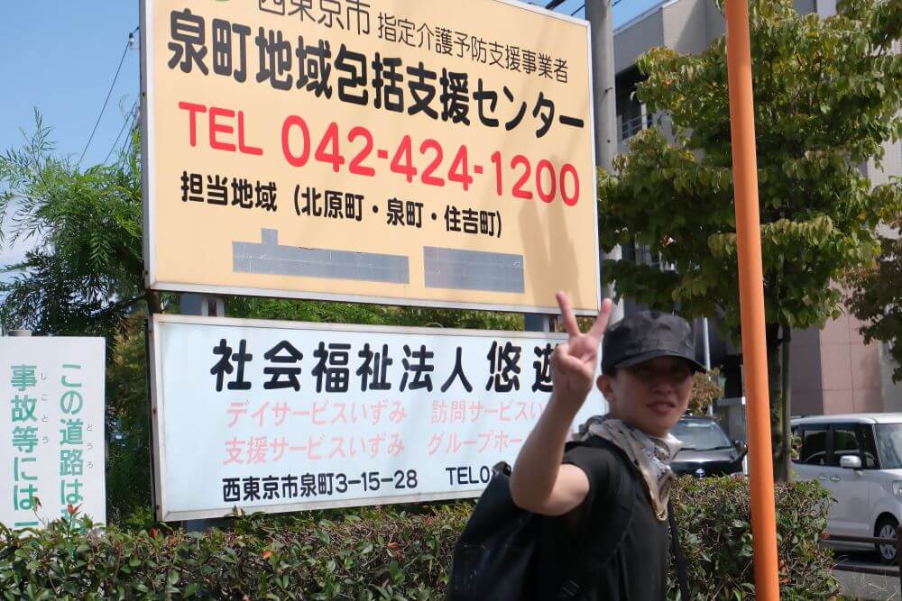 社会福祉法人　悠游の看板の前にピースするダンスの先生
